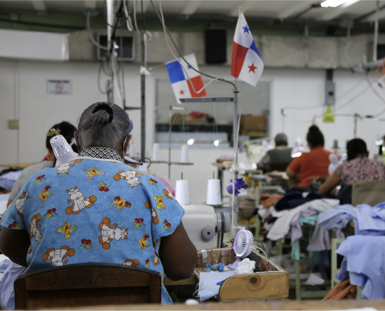 Costurera de espaldas en el taller de confección, con banderas de Panamá colgadas sobre las máquinas de coser