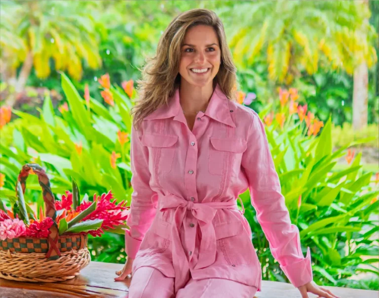 Mujer sonriente con camisero rosado de lino con bolsillos de guayabera y lazo a la cintura, sentada junto a una canasta de flores tropicales