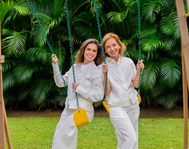 Dos mujeres sonrientes con blusas y pantalones de lino blanco, sentadas en columpios frente a un fondo de palmeras tropicales